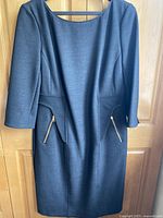 Charcoal grey fitted wool dress with 3/4 length sleeves, gold zipper and leather trim detail, hanging on hanger in front of wooden door