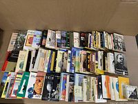 Photo showing a large collection of paperback books spread out in a box, titles and spines visible with a mixture of colors and conditions.
