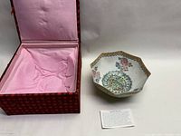 Hexagonal porcelain bowl placed next to red patterned fabric-covered box with pink satin interior and an informational card.