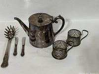 Photo of Geislingen silver tone teapot along with two tea glass holders, two butter spreaders, and one serving spoon showing tarnish and usage marks.