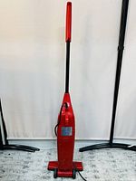 Red Simpli Stik stick vacuum cleaner standing upright against a white background, showing the back with power cord and red handle.