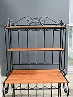 Front view of baker's rack showing three walnut finish wood shelves mounted on black metal frame with decorative metal scrollwork and hooks under the top shelf.