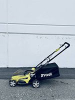Side view of Ryobi battery powered lawn mower showing yellow body, black and yellow folding handle, and black fabric collection bag with 'RYOBI' label.