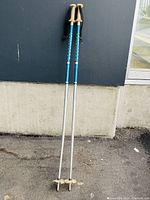 Pair of blue and silver Rossignol ski poles with tan handles and black wrist straps leaning against a wall outdoors.