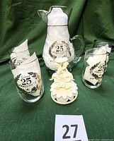 Overall view of the 25th Anniversary glass pitcher and four glasses displaying the embossed wreath and silver text, with napkins inside and the plastic cake topper in front.