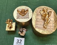 All items displayed on green fabric backdrop with inventory card #32 visible; shows sizes, shapes, and angel embellishments of gold boxes alongside the Willow Tree figurine.