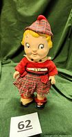 Front view of the vintage Campbell Soup Kid doll in Scottish tartan outfit including hat, red shirt, kilt, socks, and shoes on a green fabric background.