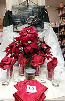 Full lot setup showing poinsettia centerpiece, eight glasses with burgundy napkins inside, candle and Thomas Kinkade tapestry hanging above.