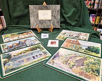 Six laminated table mats featuring painted illustrations of English inns arranged in a fan-like layout on green fabric, with the original box standing at the back.