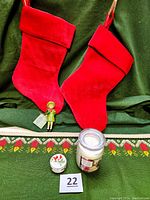 Two red velvet Christmas stockings, vintage girl skating ornament, ceramic cardinal trinket box, and Ashland candle are shown placed over a woven table runner with candle motifs on a dark green fabric background.