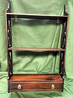 Front view of the dark brown wooden 3-tier wall shelf showing the drawer with brass-colored pulls and decorative side cutwork.