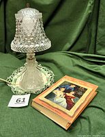 Full view of the lamp with hobnail patterned glass shade on a green fabric background, showing the lamp and a book beside it.