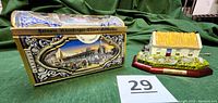 Photo of the ceramic Irish cottage music box with a wood base next to the Nuremberg dome-shaped decorative tin music box, showcasing both items together on a green cloth background with lot number.