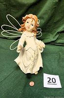 Front view of a white resin angel figurine with wire wings, holding a basket of flowers, standing on green fabric background with a small coin for scale.
