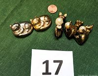 Five tiny cat figurines displayed on green fabric with a penny for scale and lot number card '17'. Includes two glazed ceramic sleeping cats in gold and silver tones, and three metal Siamese cats holding faux pearls with pink rhinestone eyes.