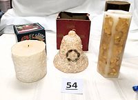 Three vintage candles arranged side by side showing textures and size differences; Travertine candle rectangular with gold foil decoration, bell-shaped candle in red and brown box, frosted pillar candle near its box with label showing a similar bumpy texture candle.