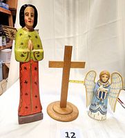 Photo shows three wooden religious-themed items: a tall wooden angel with green and red paint and praying hands, a smooth oak cross on a circular base, and a smaller blue and white angel holding a dove.
