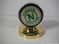 Front side of the puck displaying the Minnesota North Stars logo in protective case on gold-tone stand.