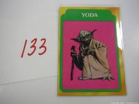 Photo of 1980 Topps Star Wars Yoda Rookie card with card number 133 next to it, showing green and yellow border with pink background featuring Yoda holding a cane.