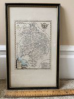 Framed antique map of the County of Warwick displayed against a baseboard and carpeted floor, showing detailed map features and scale ruler below.