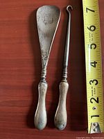 Two silver shoe tools shown side by side on a wood surface next to measuring tape, approximately 6-7 inches long, one shoe horn with broad flat scoop and one slender hooked lace puller.