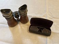 Pair of vintage binoculars with metal and brown leather casing alongside old gold-tone eyeglasses in dark velvet-lined metal case on a light fabric background.