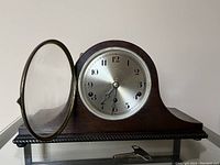 Front view of wooden mantle clock with open glass door showing silver clock face and black Arabic numerals.