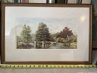 Frontal image of framed watercolour print showing Sheepwash Bridge with river, trees and cottage in natural colors, measuring approximately 24 x 16 inches.