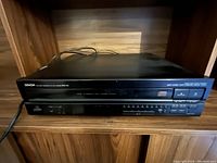 Front angled view of the Denon DCM-420 5-disc CD player on a wooden shelf showing power cord and front panel buttons.