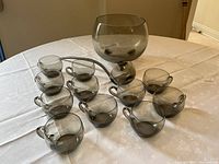 Photo showing eleven smoke grey glass punch glasses with handles, one large punch bowl, and one ladle placed on a white tablecloth.
