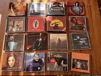 Top view of 16 music CD jewel cases arranged in a 4x4 grid on a wooden surface, showing a variety of album covers from different artists and music genres.