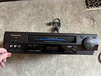 Front angle view of Panasonic PV-4554 VHS VCR showing front panel tape slot, AV inputs, and control dial with power cord wrapped behind.