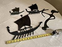 Overview photo showing all three iron Viking ship wall decorations lined up with measuring tape for scale, alongside the Greek bronze statue.