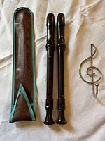 Two Yamaha black plastic recorders, a brown leather case with turquoise trim, and a silver treble clef music note-shaped hook on fabric background