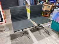 Two black fabric chairs with aluminum four-legged bases, shown together on a concrete floor in a warehouse setting.