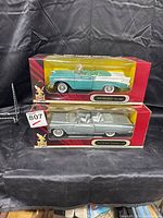Two unopened diecast car models in their original red and yellow boxes with clear plastic windows showing the cars inside; one turquoise/white 1956 Chevrolet Bel Air and one silver 1959 Buick Electra 225.