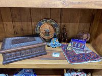 Full view showing all lot items on wooden shelf including hand carved wooden box, brass Buddha statues, ceramic meditation figure, African resin figure, copper camel plate, blue enamel trinket box, heart-shaped copper ashtray and fabric mats