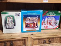Three boxed ceramic Christmas village buildings displayed side by side on wooden surface.