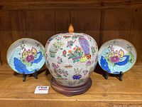 Front view of the Chinese crackle glaze jar with colorful hand-painted designs and two Japanese plates displayed on either side showing flower motifs and brass rims.