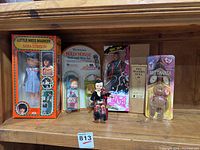 Wide shot of all dolls and toys displayed on wooden shelf showing all six items with boxes or original packaging, including Little Miss Marker, Holly Hobbie, NKOTB Jordan doll, Peg Wooden Doll, Ty Britannia Beanie Baby and small sitting doll.