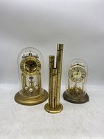 Photo shows 2 mechanical mantle clocks and 1 brass cylindrical coin organizer under glass domes on a white surface.