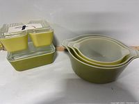 Photo shows three nested Pyrex mixing bowls with gradient olive green to white finish on the right and three stacked rectangular Pyrex refrigerator dishes with clear glass lids in olive green and yellow on the left.