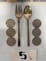 Top-down image showing the sterling fork and spoon positioned vertically with six oversized dollar coins arranged on either side.