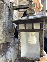 Close-up of outdoor wall light fixture attached to stone wall, showing greenish-dark grey finish and frosted glass panel with spiral compact fluorescent bulb inside.