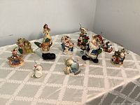 Overview of 11 assorted Christmas ornaments mostly ceramic or resin on a patterned white tablecloth all visible with various holiday characters including Santa, angels, snowmen, and elves.