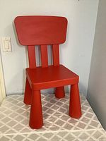 Front and side angle view of the bright red child-sized IKEA chair, showing the seat, backrest with three vertical slats, and wide tapered legs.