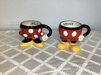 Front view of two ceramic mugs styled as Minnie and Mickey Mouse characters with distinct colors and signature details on rims.