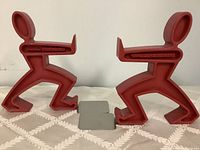 Pair of red plastic bookends shaped as stylized human figures in dynamic poses, facing each other to hold books.