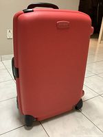 Closed bright red hard shell suitcase with four wheels and black handle, sitting on tiled floor.