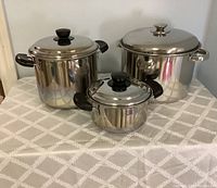 Three stainless steel pots with lids displayed on a patterned cloth surface